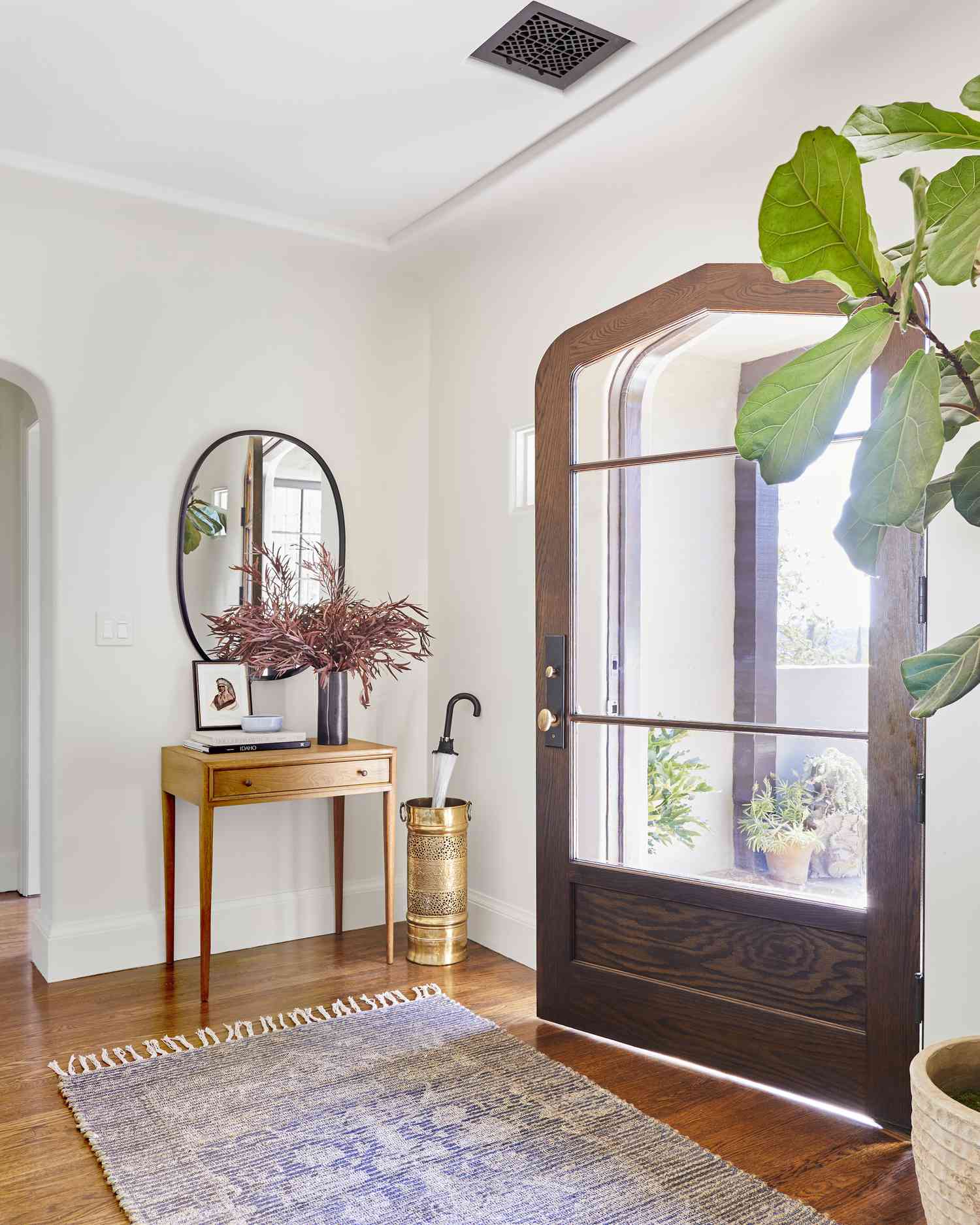 A large mostly glass door opens to a small light wood side table above which hangs a circular mirror 