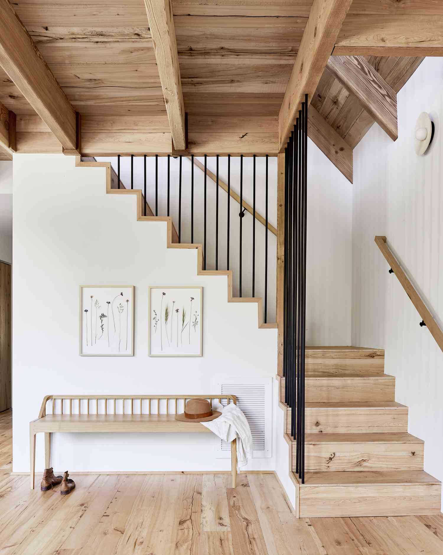 A white room has rustic style uncreated wood timbering for a roof and wood stairs. The entryway has a small bench and two wood framed art pieces of pressed flowers.