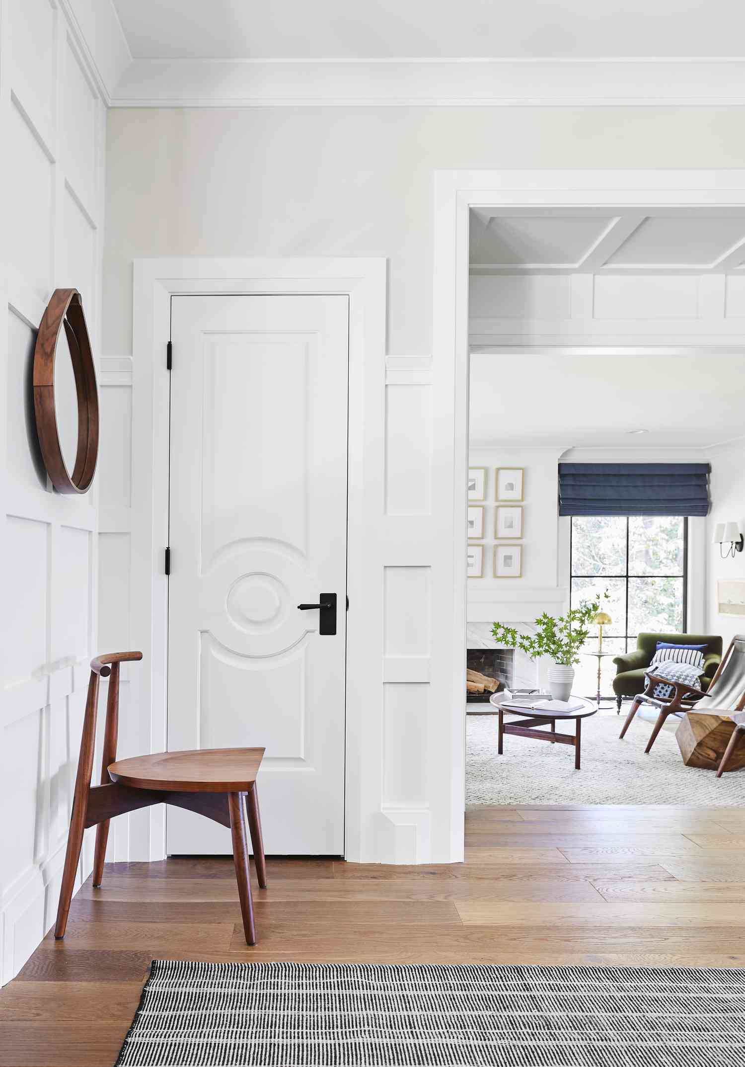 A white paneled wall near the entryway is covered in an irregularly shaped wood framed mirror below which sits a modern wood chair