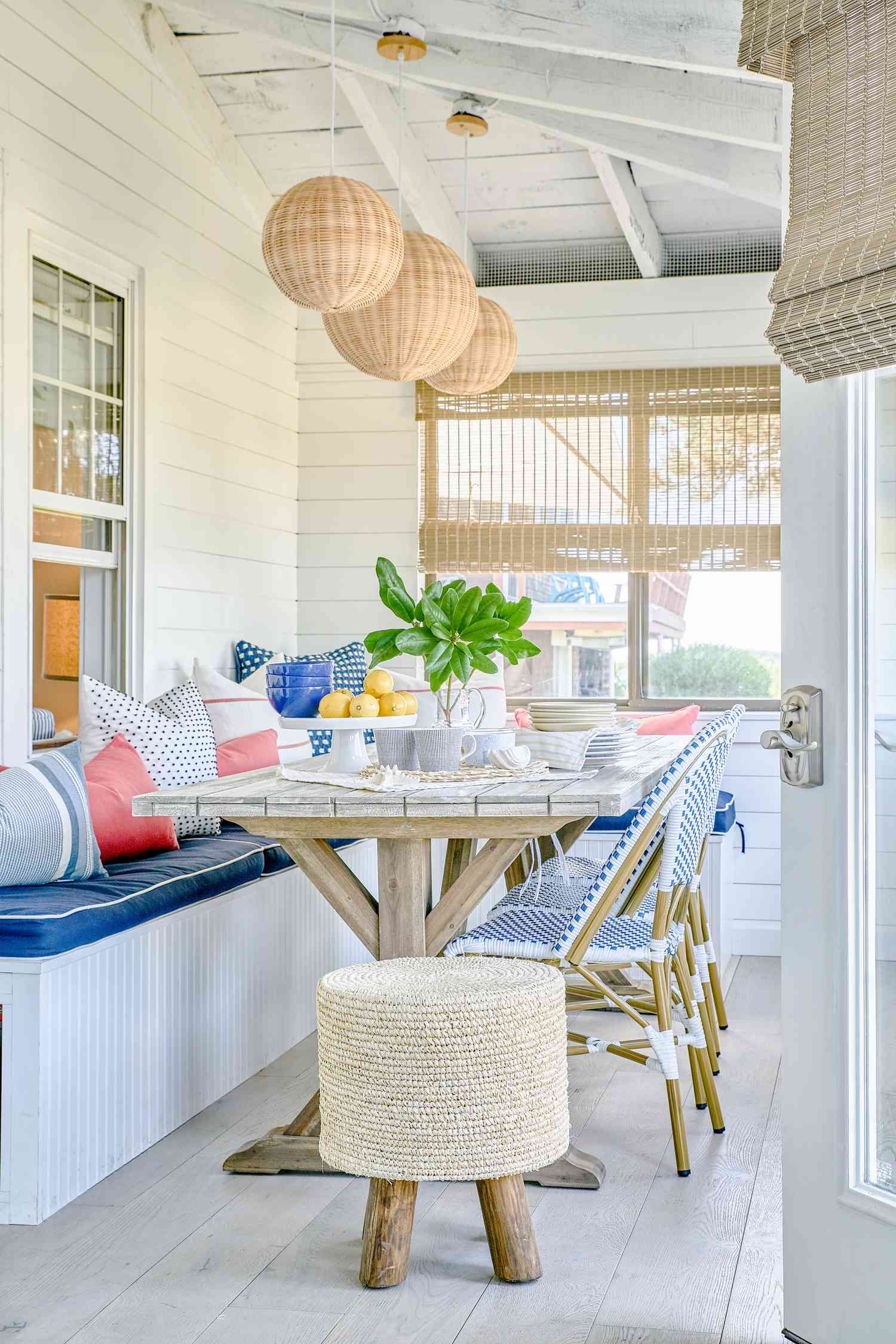 Small sunroom with a built-in dining bench around a wood table with blue and white chairs and rattan pendant lights hanging from the ceiling