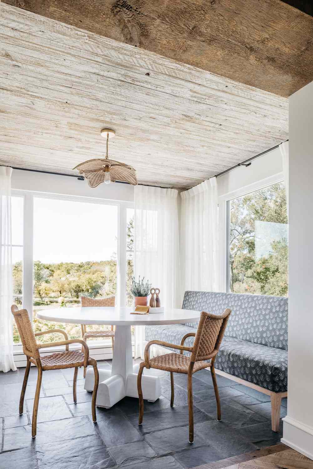 Sunroom with white curtain panels and wood ceiling with a round white table, upholstered settee and wicker chairs